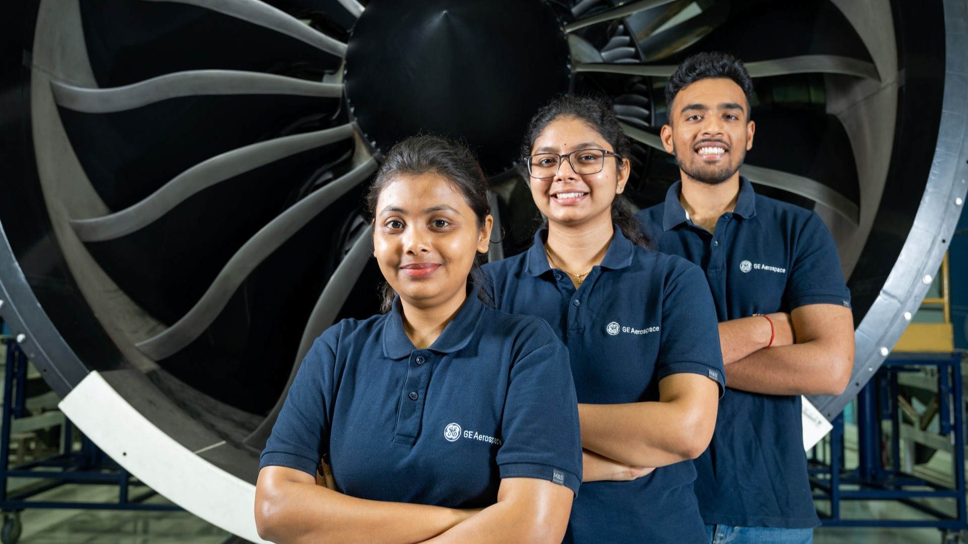 Three engineers in blue polos stacked standing in front of an engine