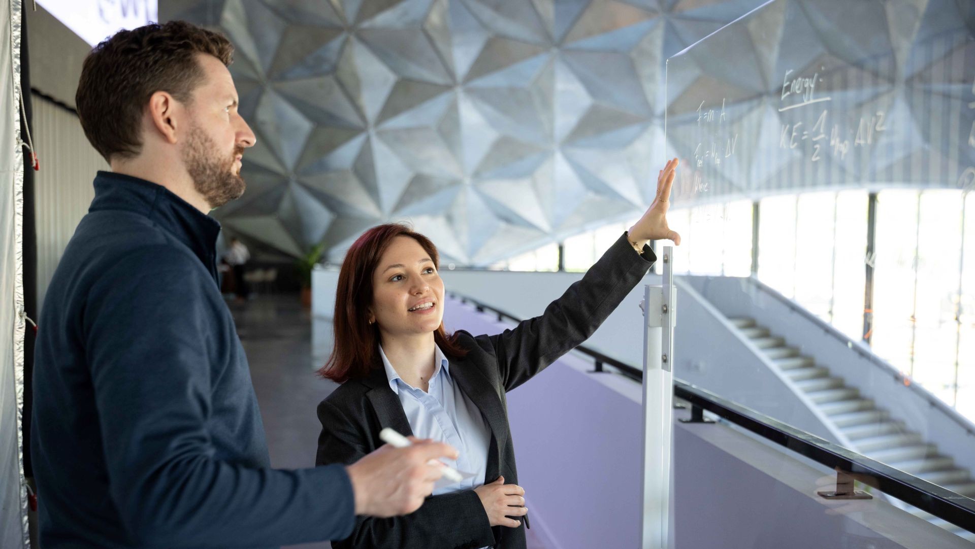 Woman gesturing to a clear board of equations with a man by her side